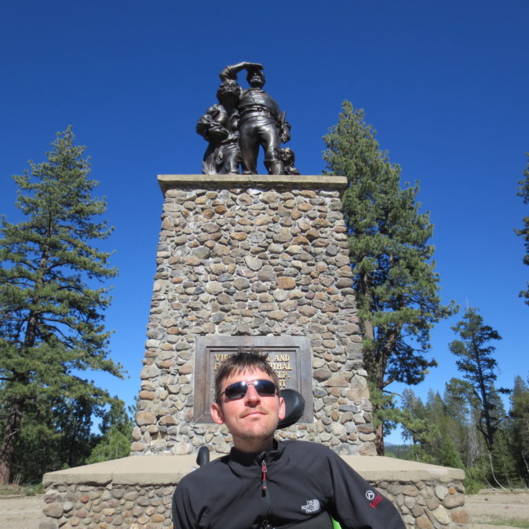 Brian, wearing sunglasses and a black jacket, sitting in his wheelchair in front of a statue of a pioneer family commemorating the Donner party tragedy.